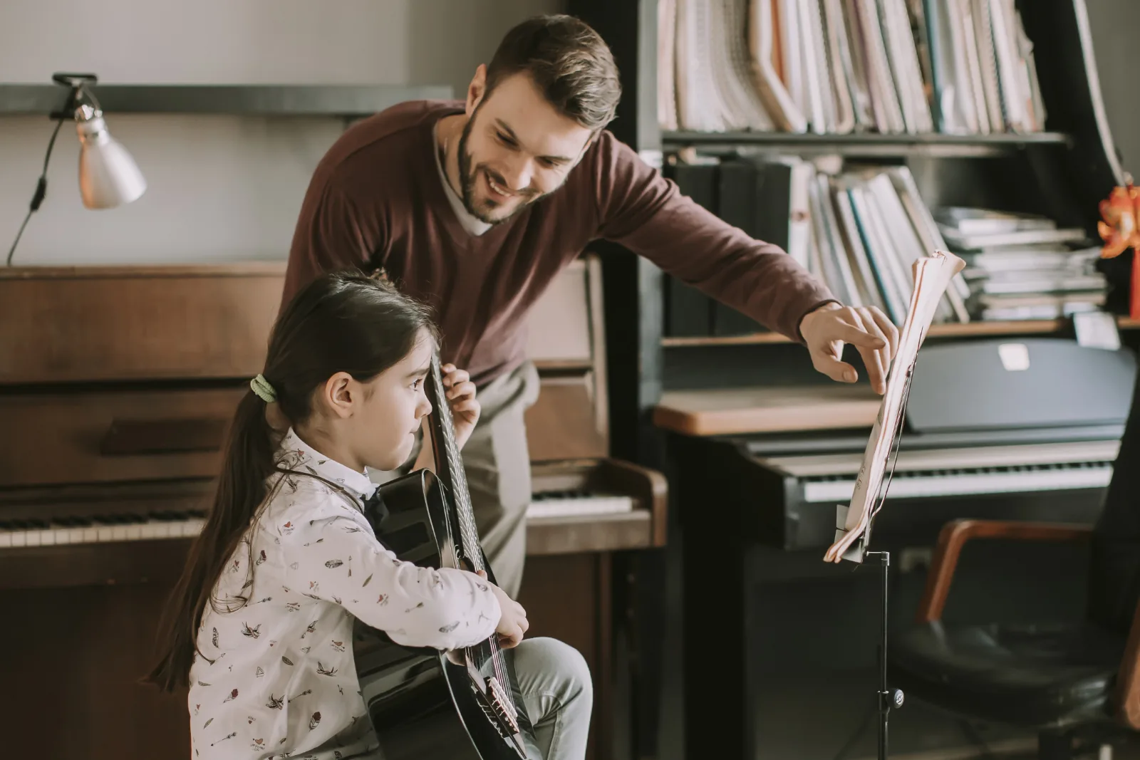 Music teacher guiding a young student playing classical guitar from a music stand.