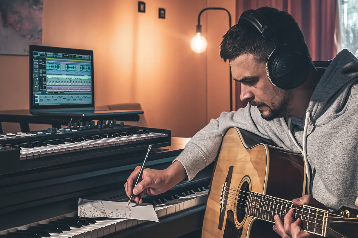 A musician transcribing at a piano with a guitar and a DAW.
