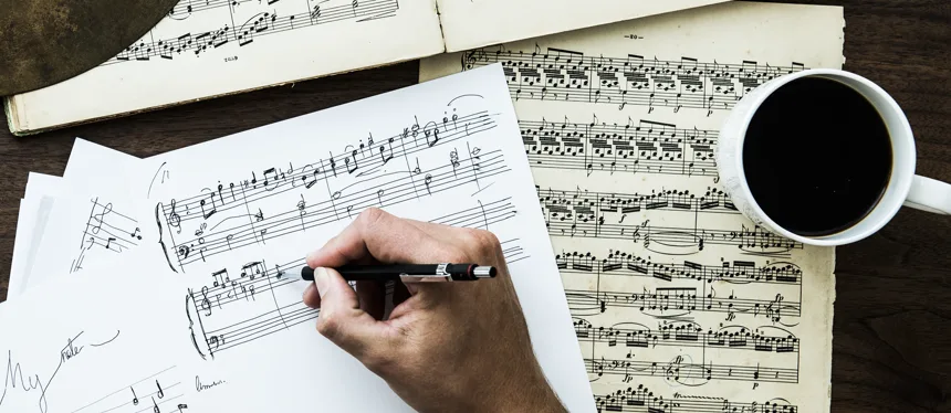 A hand writing music notation on manuscript paper beside aged sheet music and a cup of coffee on a wooden desk.