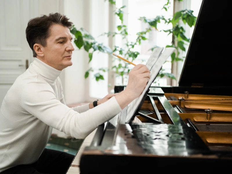 Composer seated at a grand piano, annotating a score in pencil