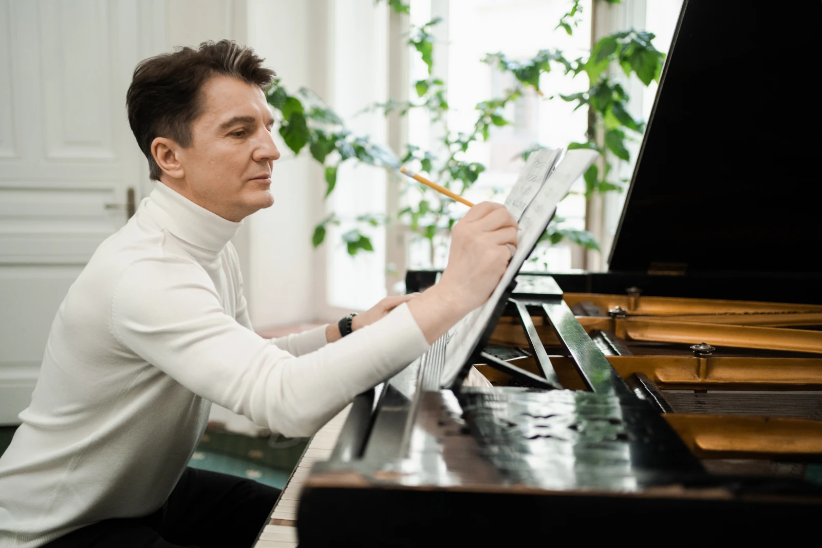 A composer seated at a grand piano, annotating a score in pencil.