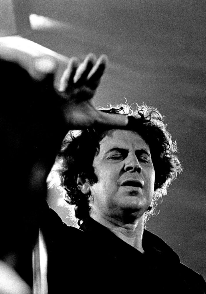 Mikis Theodorakis conducting, eyes closed, one hand raised
